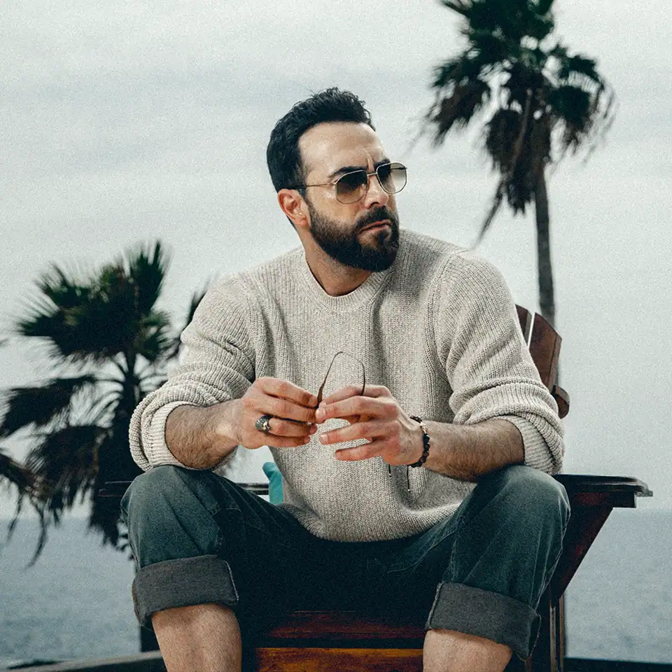 Lebanese actor Carlos Azar in a casual, outdoor portrait, wearing sunglasses and a cream sweater, photographed by Jean-Claude Dib.