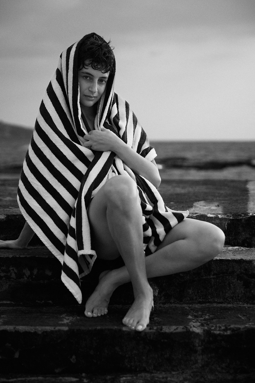 Black and white editorial portrait of actress Isabelle Zeghondi wrapped in a striped towel on a coastal ledge in Lebanon, shot by film director and fashion photographer Jean-Claude Dib